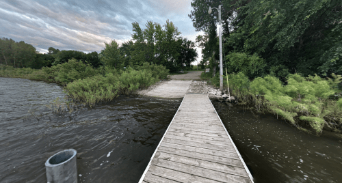 A wooden dock extends over water, surrounded by lush greenery and trees under a cloudy sky.