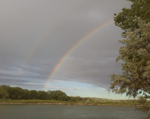 A double rainbow arcs over a river, framed by dark clouds and green trees along the shore.