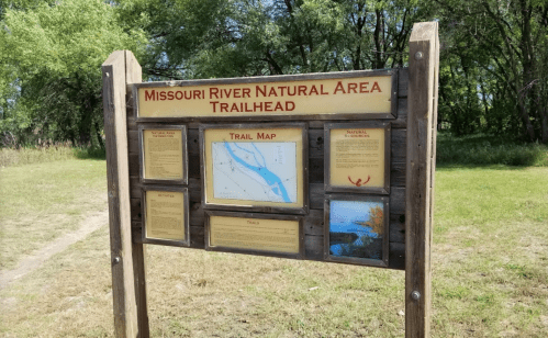 Sign for Missouri River Natural Area Trailhead, featuring a trail map and information about the area. Green landscape in background.