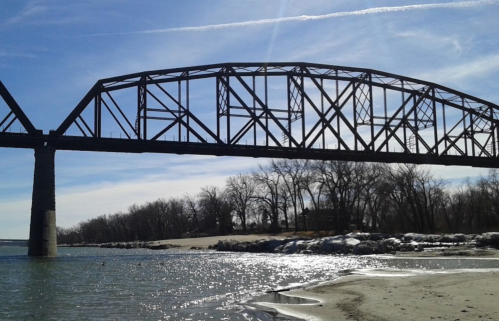A large metal bridge spans over a river, with sandy banks and trees in the background under a clear blue sky.