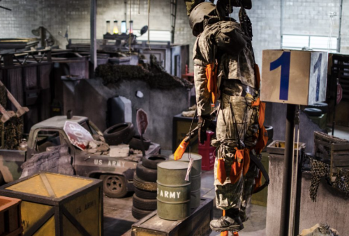 A military training facility with a mannequin in gear suspended above barrels and crates, surrounded by vehicles and obstacles.