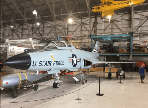 A U.S. Air Force fighter jet displayed in a hangar, with various aircraft parts and equipment in the background.