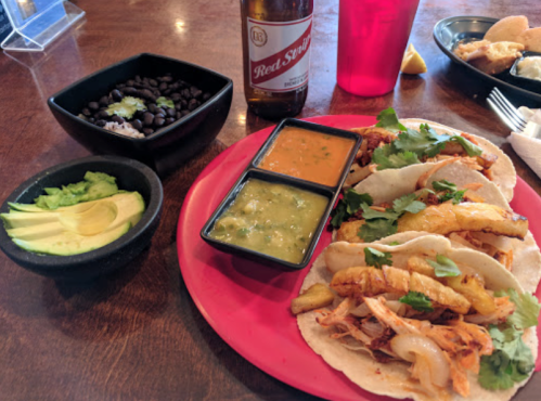 A plate of tacos with chicken and toppings, served with black beans, avocado slices, and two sauces, alongside a drink.