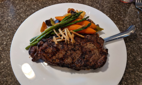 A grilled steak served on a white plate with asparagus, carrots, and crispy potato strips on the side.