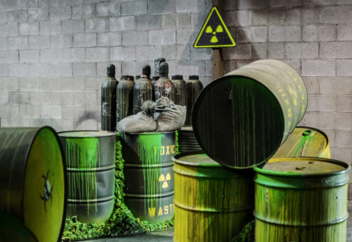 A dimly lit industrial space with green toxic waste barrels, a warning sign, and gas cylinders in the background.