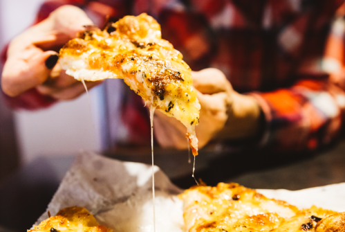A person holding a slice of cheesy pizza, with melted cheese stretching from the slice to the pizza tray.