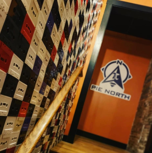 A colorful wall of cassette tapes leads to a doorway with a "Pie North" logo on an orange background.