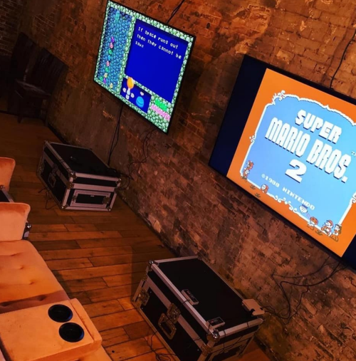 Two TVs on a brick wall display classic video games, with seating and equipment cases in a cozy gaming space.