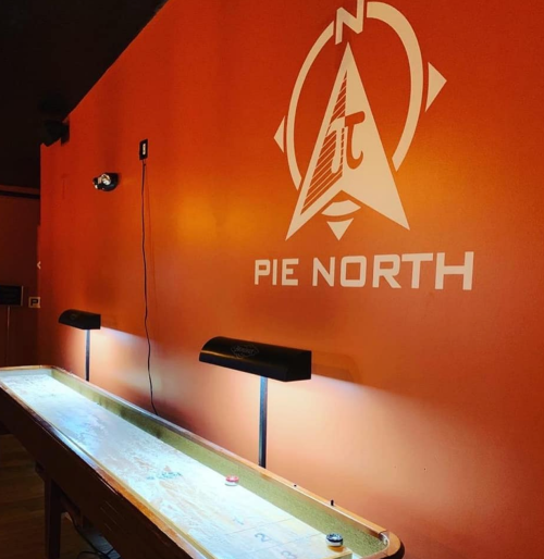 A wooden shuffleboard table under two lamps, with a bright orange wall featuring the "Pie North" logo.