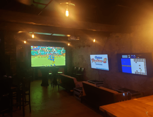 A cozy gaming lounge with two large screens displaying retro video games, dim lighting, and seating areas.