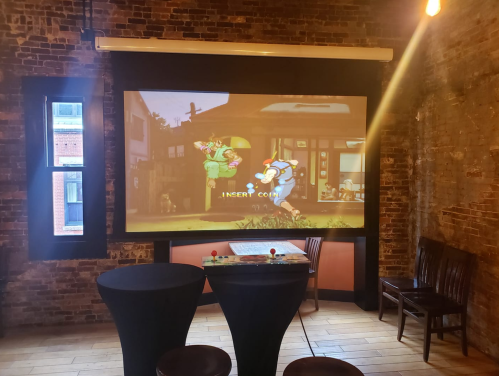 A retro arcade setup with a large screen displaying a game, two tables, and wooden chairs in a brick-walled room.