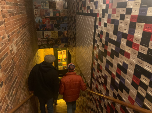 Two people descend a staircase lined with colorful cassette tape artwork, leading to a warmly lit doorway below.
