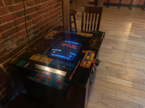 A vintage arcade game table with a glass top, featuring colorful controls and a brick wall backdrop.