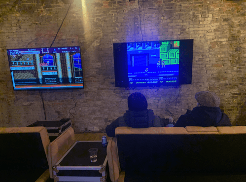 Two people sit on a couch, playing retro video games on two wall-mounted screens in a cozy, brick-walled room.