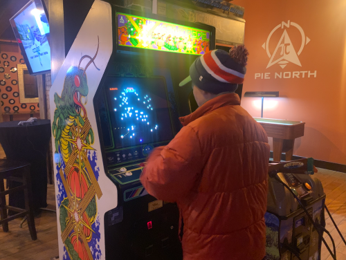 A person in an orange jacket plays a retro arcade game in a cozy bar setting.