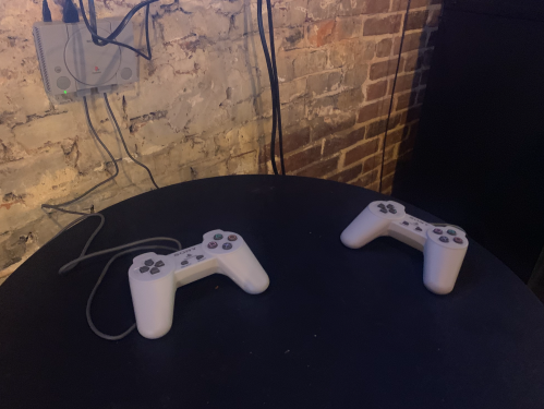 Two white game controllers on a black table, with a PlayStation console mounted on a brick wall in the background.