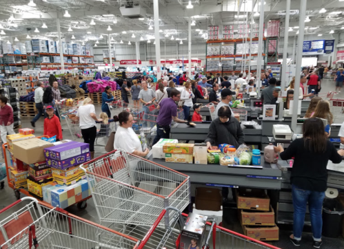 A busy Costco checkout area with many shoppers and shopping carts filled with various products.