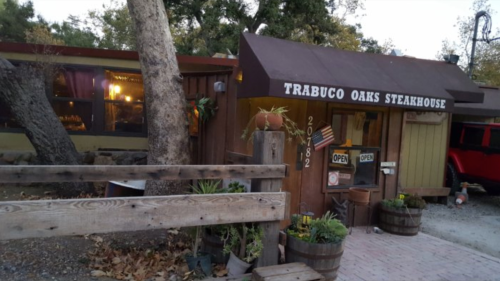 A rustic steakhouse with a wooden exterior, open sign, and potted plants, surrounded by trees and a red vehicle.