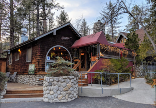 A cozy wooden cabin with a stone entrance, surrounded by trees and festive lights, set in a serene outdoor environment.