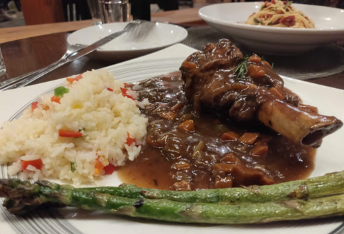 A plate featuring a lamb shank in sauce, rice with vegetables, and grilled asparagus.
