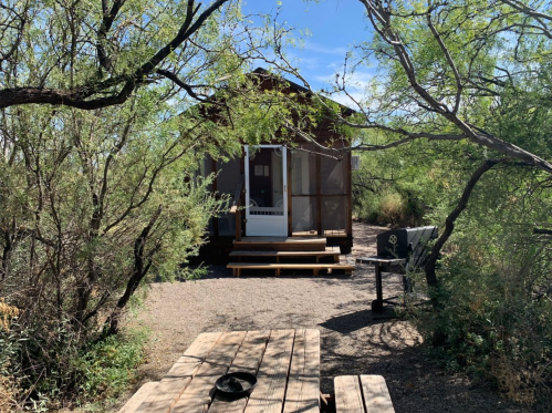 A small cabin surrounded by greenery, with a picnic table and grill in the foreground under a clear blue sky.