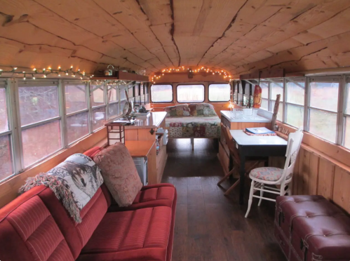 Cozy interior of a converted bus with wooden walls, string lights, a bed, seating area, and a dining table.