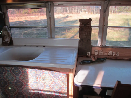 Interior of a bus converted into a living space, featuring a sink, table, and owl decorations by the window.