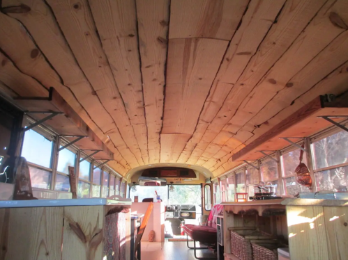 Interior of a converted bus with wooden ceiling, large windows, and cozy seating areas. Natural light fills the space.