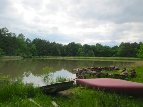 A serene lake surrounded by greenery, with a canoe and a stone fire pit in the foreground under a cloudy sky.