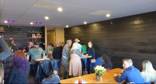 A lively gathering in a cozy venue with people socializing around wooden tables and a dark wood-paneled wall.