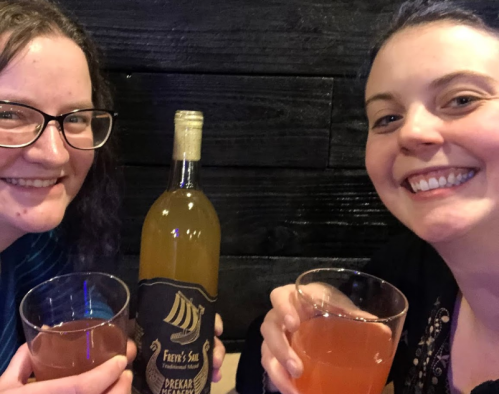Two smiling friends hold drinks and pose with a bottle of wine on a dark wooden background.