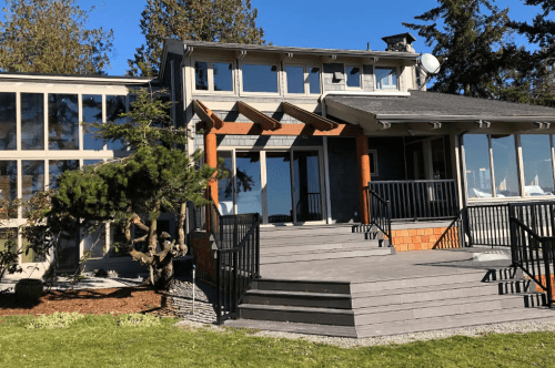 A modern house with large windows, a wooden deck, and a landscaped yard surrounded by trees.