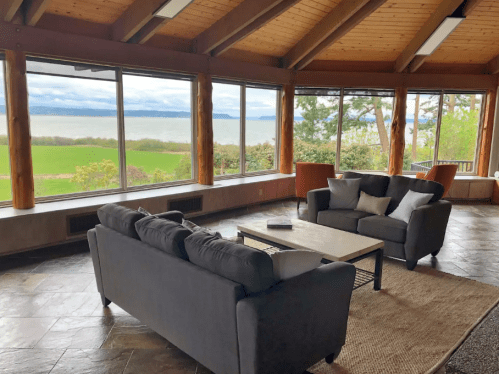 Cozy living room with large windows overlooking a scenic lake and green landscape. Modern furniture and natural light.