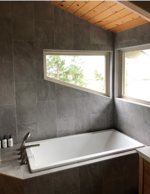 A modern bathroom featuring a freestanding tub, gray tiled walls, and large windows with a view.