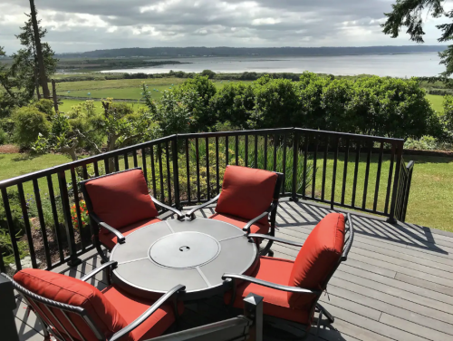 A deck with four red chairs around a circular table, overlooking a scenic view of green fields and a body of water.