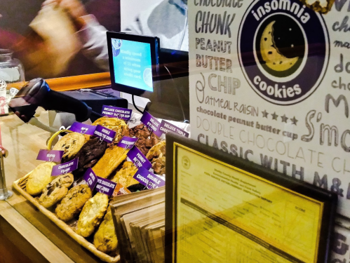 A display of various cookies at Insomnia Cookies, featuring labels for different flavors and a menu in the background.