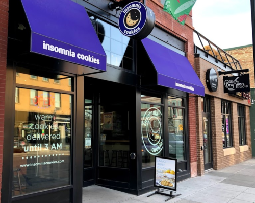 Exterior of Insomnia Cookies shop with purple awnings, showcasing a sign for warm cookie delivery until 3 AM.