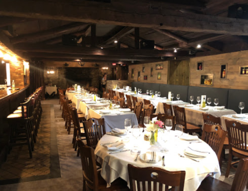 A cozy restaurant interior with wooden tables, chairs, and a bar, set for dining with flowers and glassware on the tables.