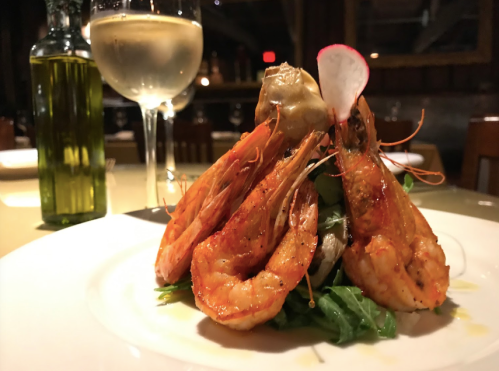 A plate of grilled shrimp on a bed of greens, accompanied by a glass of white wine and a bottle of olive oil.