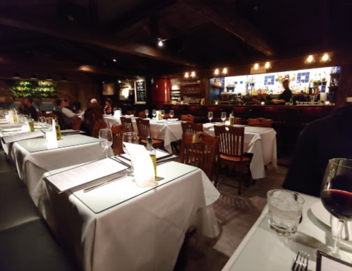 A dimly lit restaurant with white tablecloths, wooden chairs, and a bar in the background, creating an intimate atmosphere.