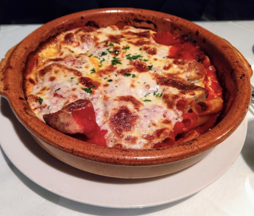 A baked pasta dish with melted cheese, tomato sauce, and herbs in a clay bowl, served on a white plate.