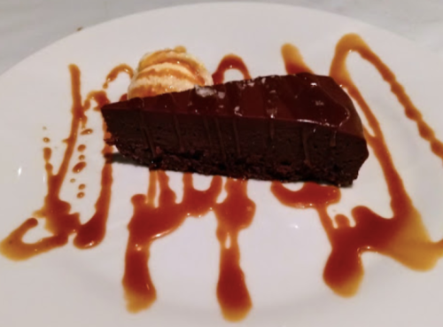 A slice of rich chocolate cake drizzled with caramel sauce, accompanied by a scoop of vanilla ice cream.