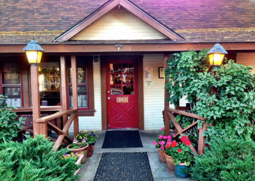 A charming restaurant entrance with a red door, surrounded by greenery and colorful flower pots, with an "Open" sign.