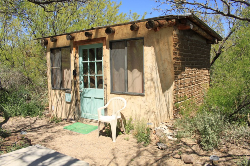A small, rustic adobe cabin with a turquoise door, surrounded by desert vegetation and a white chair outside.