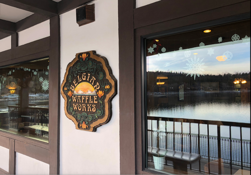 Sign for "Belgium Waffle Works" on a building, with a view of a lake and winter decorations in the windows.
