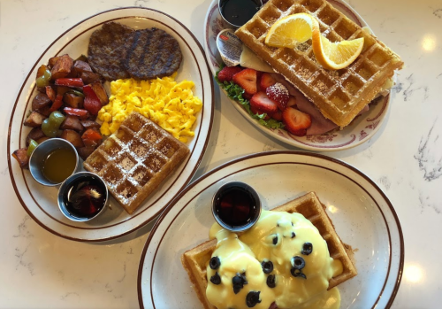Two plates of breakfast featuring waffles, sausage, scrambled eggs, potatoes, and fresh fruit with syrup on the side.