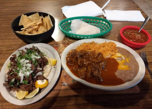 A plate of beef with onions and sauce, rice, refried beans, tortillas, chips, and salsa on a wooden table.