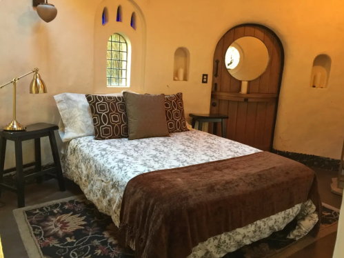 Cozy bedroom with a bed, decorative pillows, wooden door, and unique windows, featuring warm, earthy tones.