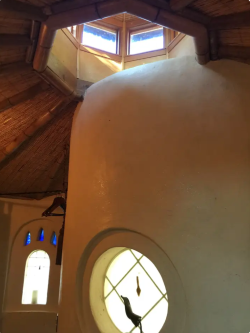 Interior of a round, straw-roofed structure with a curved wall and circular window, featuring natural light from above.