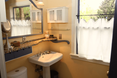 A small bathroom featuring a sink, mirror, and window with sheer curtains, decorated in warm yellow tones.